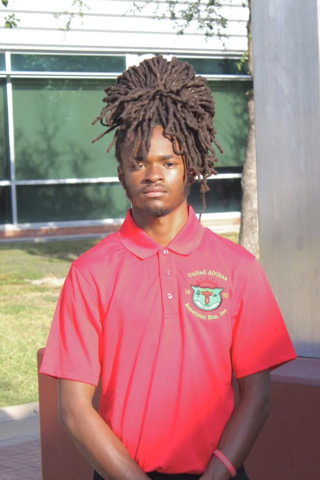 A young man with long, thick dreadlocks tied up high on his head, wearing a bright red polo shirt with a crest that reads 'United African American Men, Inc.' The background features a modern glass building and some greenery. The young man has a serious expression.