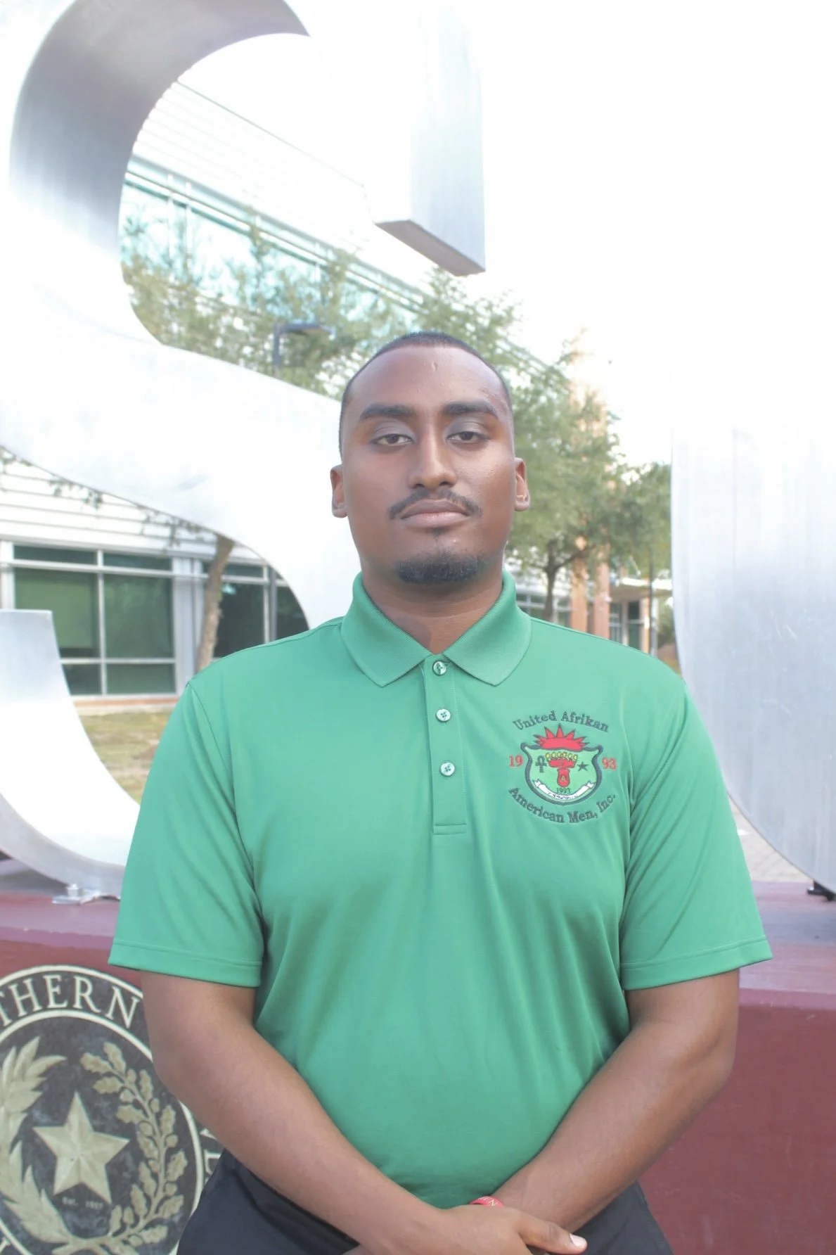 Young man standing outdoors in front of a modern building with metal sculpture, wearing a green polo shirt with a logo, looking at the camera.