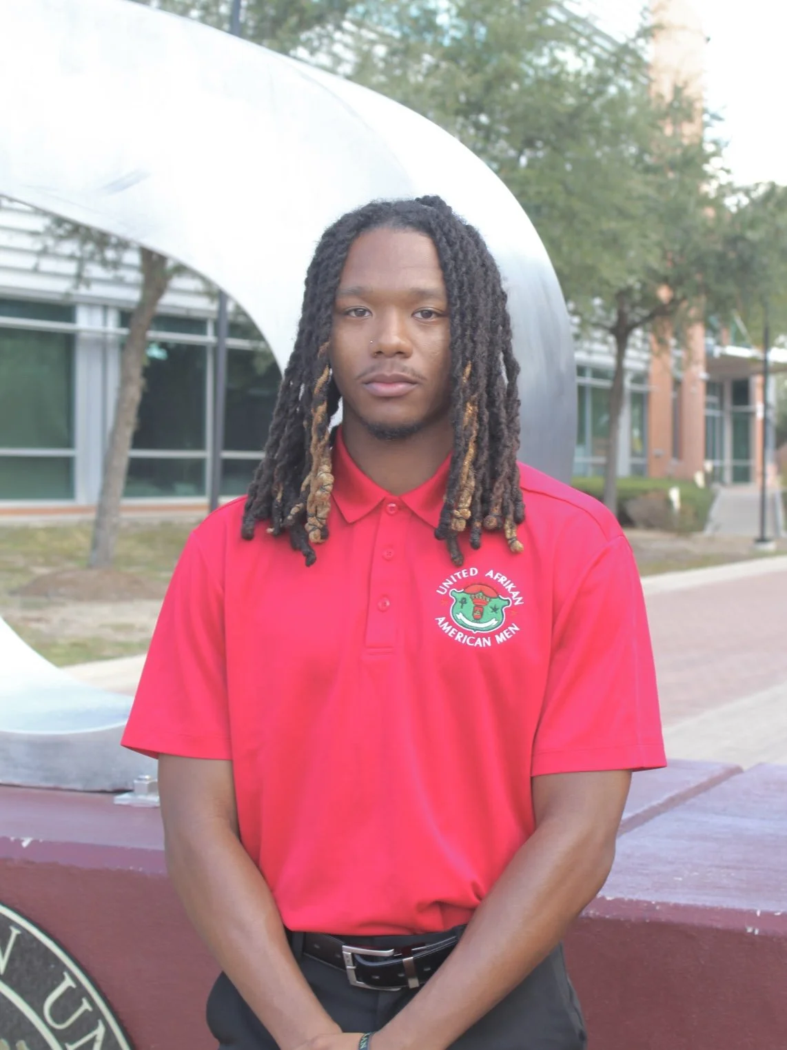 Young man with long dreadlocks wearing a red polo shirt with a United African American Men logo stands outdoors in front of a modern building and sculpture.