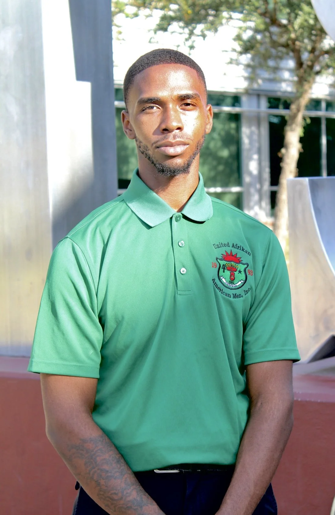 A young man with short hair and a beard wearing a green polo shirt with a crest emblem, standing outdoors near a park or campus with trees and modern buildings in the background.