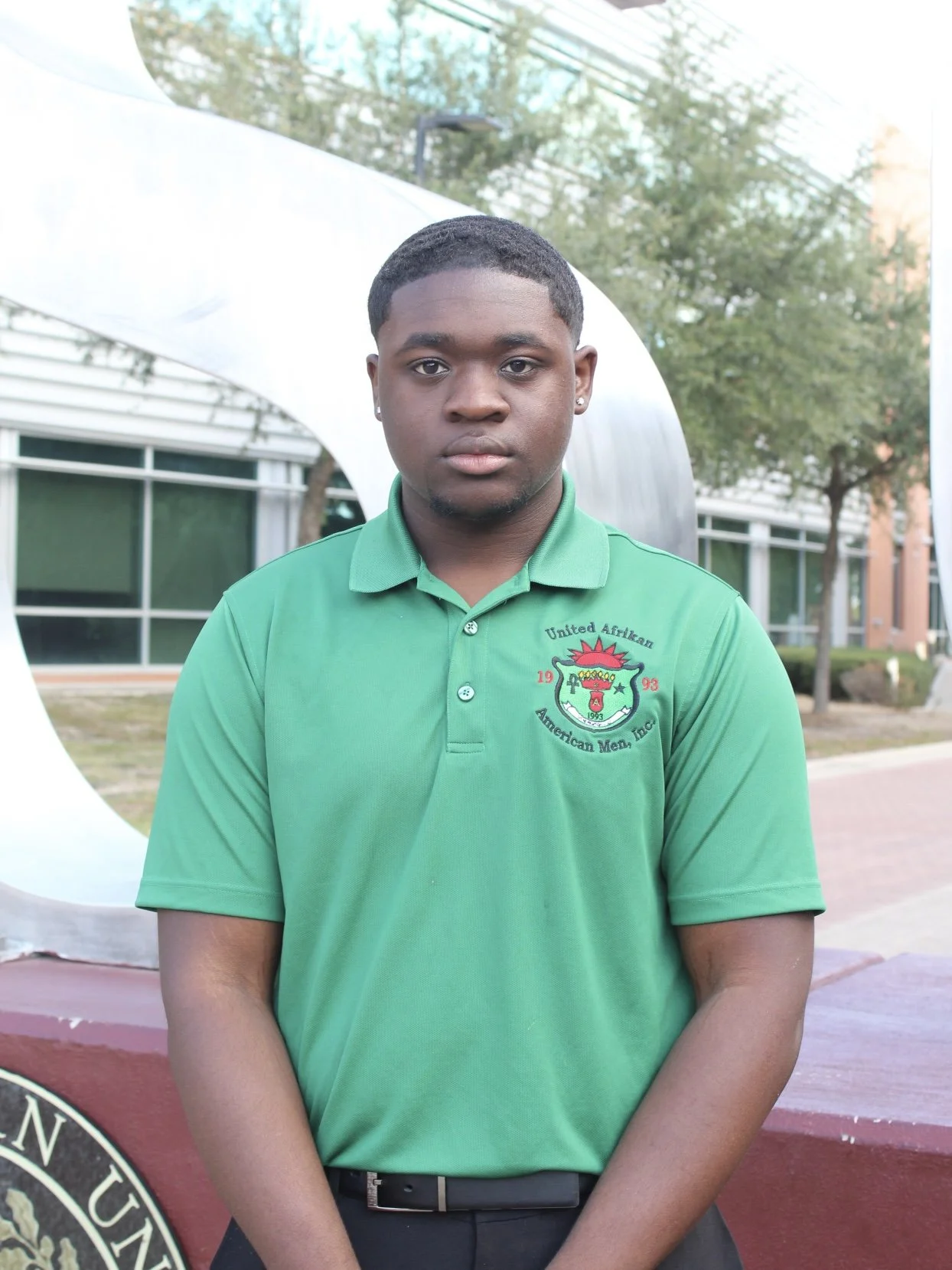 A young man with short black hair, wearing earrings and a green polo shirt with a crest, stands outdoors in front of a modern building with large glass windows and trees in the background.