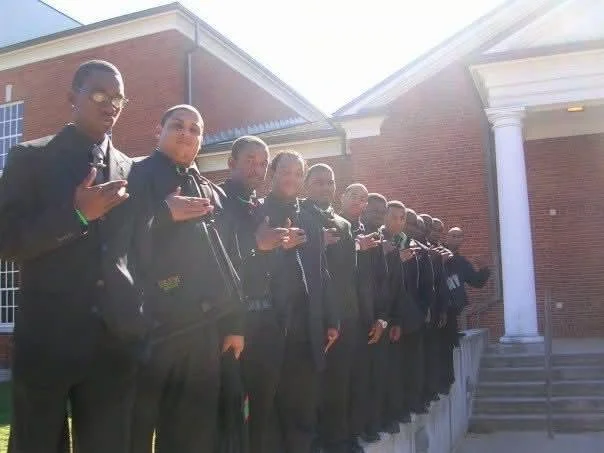 A line of young men dressed in black suits, standing outside a red brick building with white columns, holding up their hands in a salute or gesture of respect.