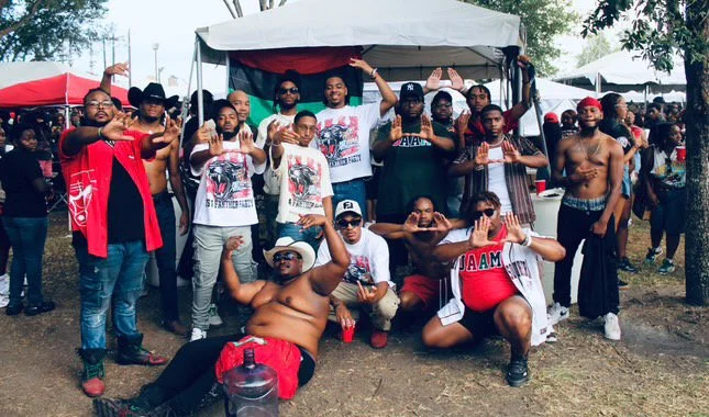 A group of men gathering outdoors, some of them shirtless, wearing casual clothing, posing for a photo in front of a canopy tent at a festival or gathering.