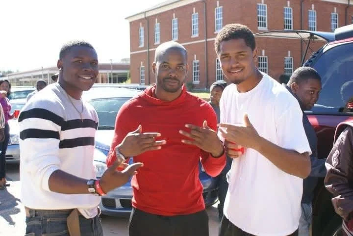 Three young men waving and smiling at the camera outdoors, with cars and a brick building in the background.