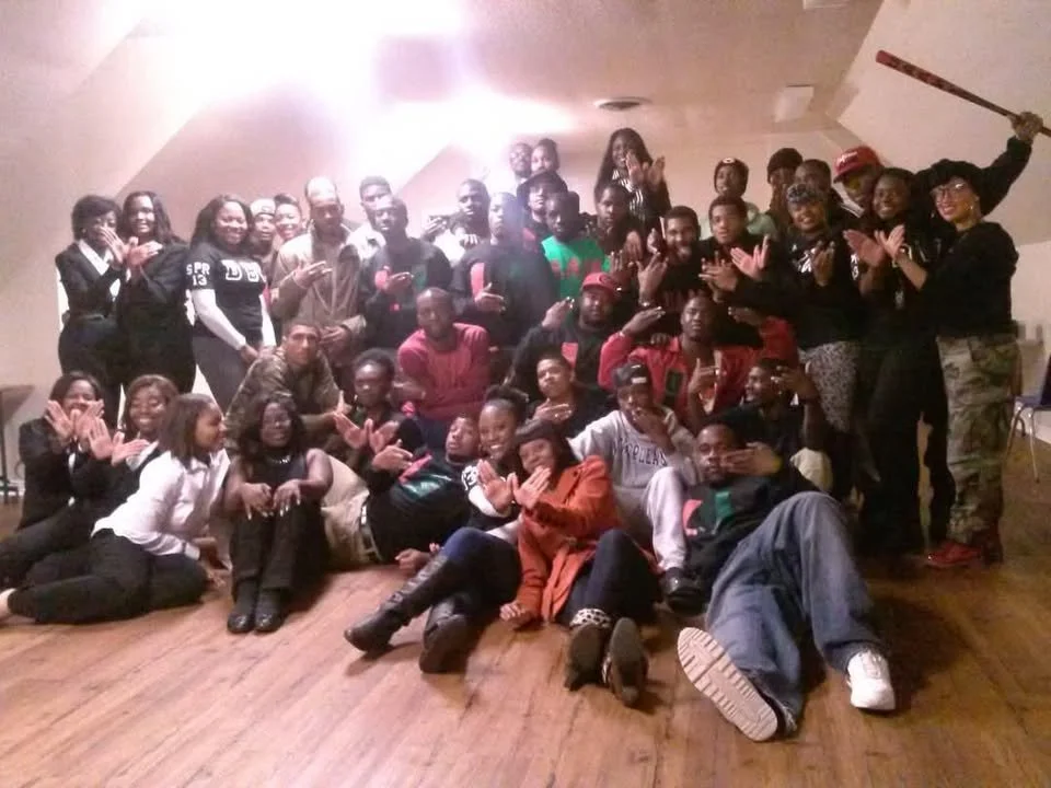 Group of people, mostly young adults, posing together in a room, some sitting on the floor and others standing behind, many smiling and making different hand gestures.