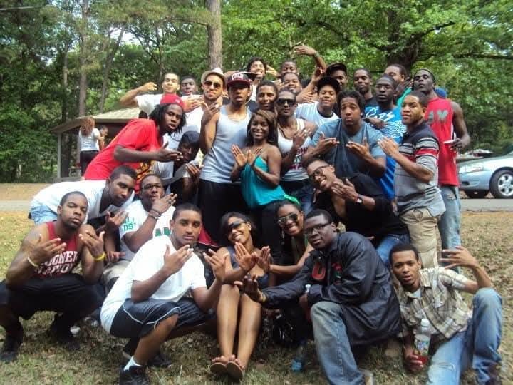 Group of young adults posing outdoors in a park with trees and cars in the background.