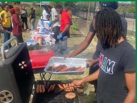 Person grilling hot dogs and sausages at an outdoor cookout with a group of people in the background.