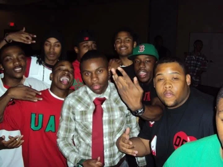 Group of young men posing for a photo indoors, some making hand gestures, wearing casual and sportswear, with a dark background.