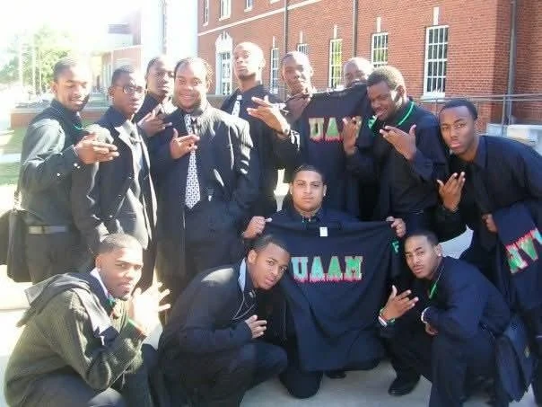 A group of young men dressed in formal attire posing outdoors, holding a black shirt with colored text, standing in front of a brick building with windows.