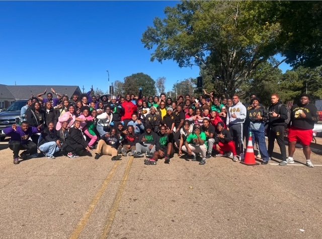 A large group of people gathered outdoors on a sunny day, posing for a group photo in a parking lot with trees in the background.