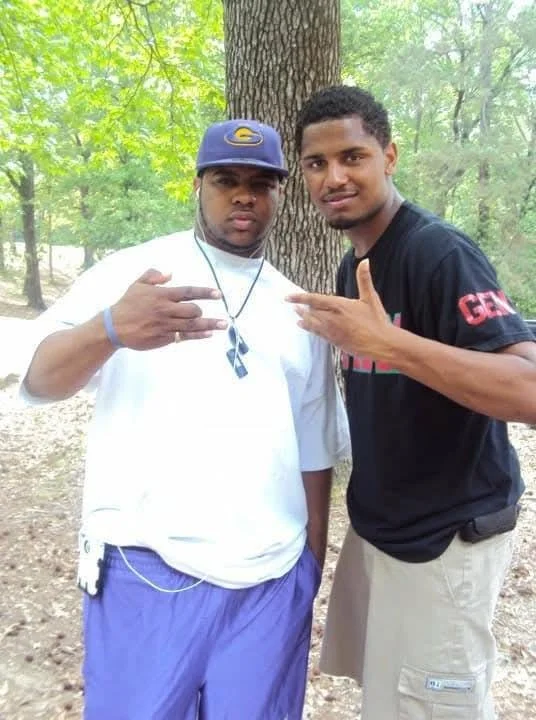 Two young men standing outdoors in a wooded area, posing for a photo. The man on the left is wearing a blue cap, white shirt, purple shorts, and has a key and a lanyard hanging from his pocket. The man on the right is wearing a black t-shirt and beig