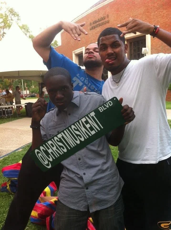 Three young men posing outdoors at a park or campus, with a brick building in the background. One man is holding a green sign with @CHRISTINSKIEN BLVD written on it, and they are making playful hand gestures and facial expressions.