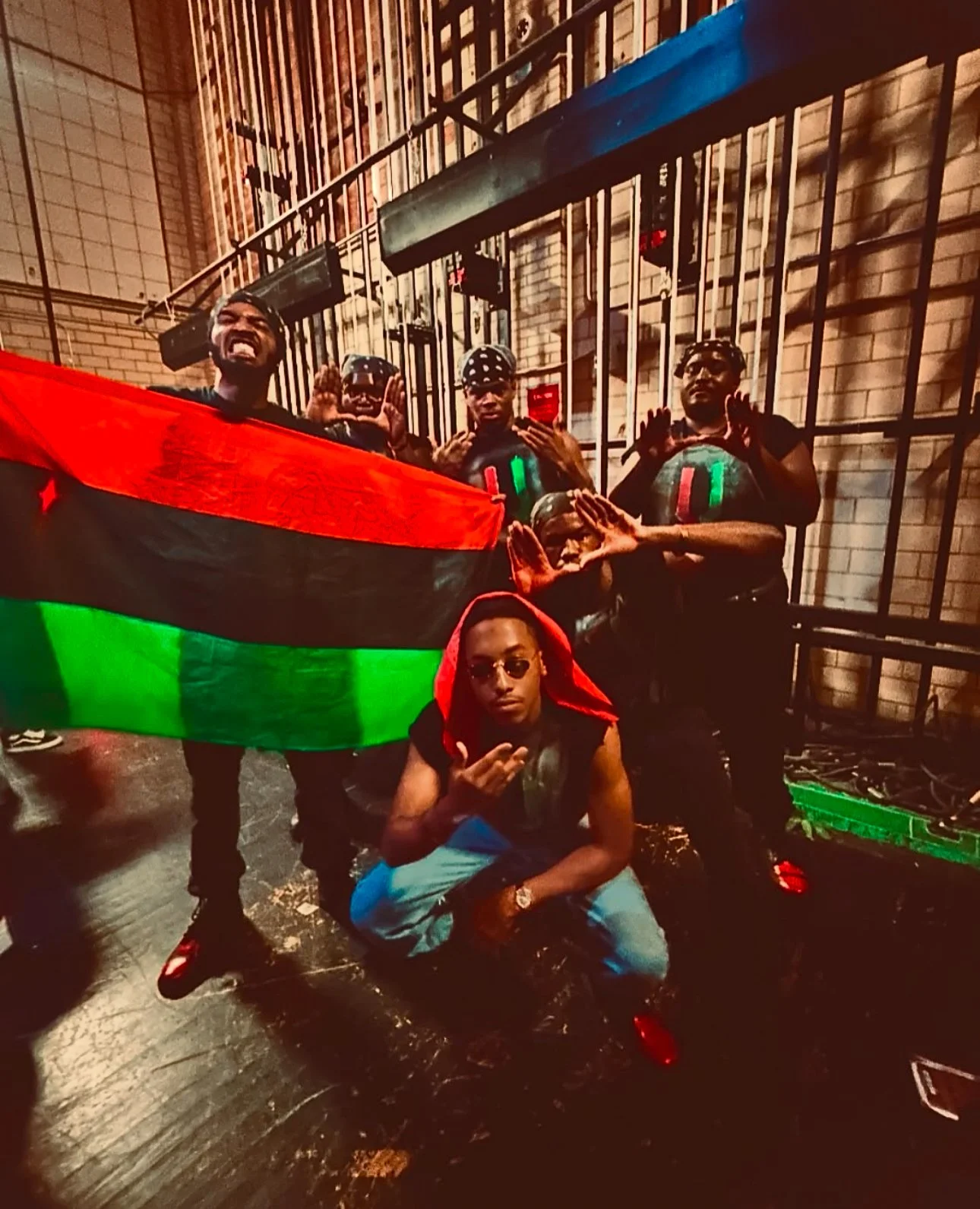 Group of six people posing with a flag and skulls in a backstage area with black bars and brick wall. Four standing with hands near their heads, one crouching in front with sunglasses and red hoodie. All are dressed in dark clothing.