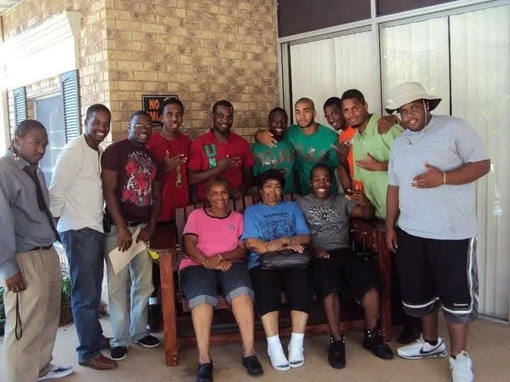 Group of people gathered indoors, posing for a photo, with some sitting on a bench and others standing behind them.