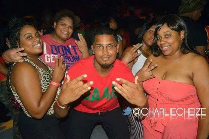 Group of five friends at a party, smiling and making hand gestures. Women wearing colorful clothing, one in leopard print, another in pink, and a man in a red shirt with green text.