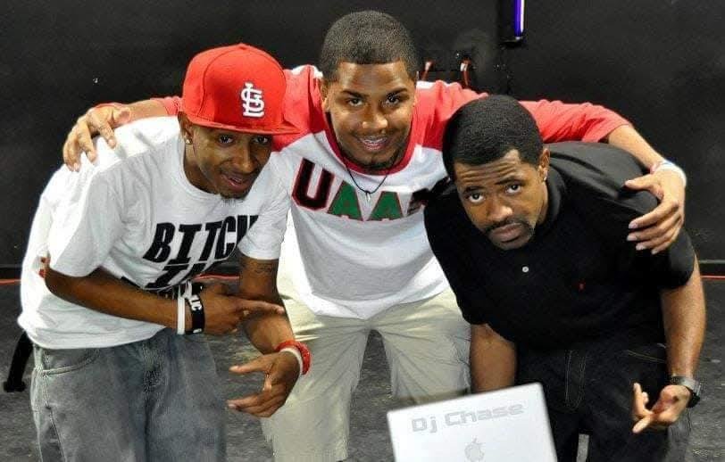 Three young men posing together, one wearing a white T-shirt with black text and a red cap, another in a white and red shirt with 'UAA' on front, and the third in a black polo shirt, with electronic equipment in front of them.