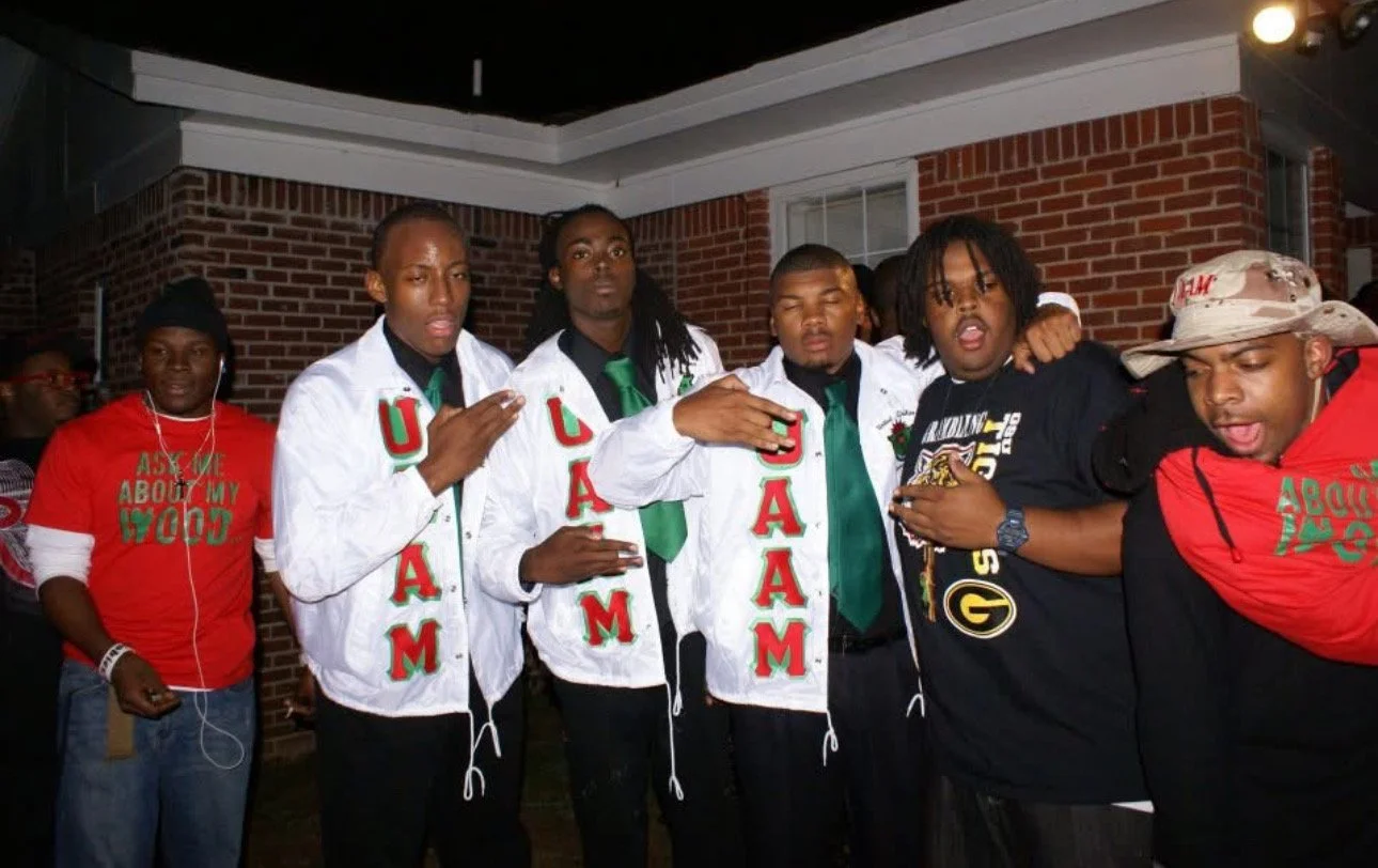 Group of young men wearing UGA and Georgia Bulldogs attire, posing together outdoors at night near a brick building, some with eyes closed or making gestures.