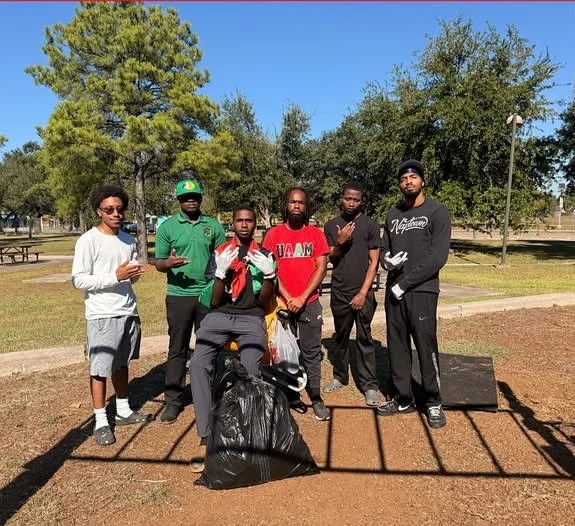 Six young men standing together at a park, some holding trash bags and gloves, during a community cleanup event.