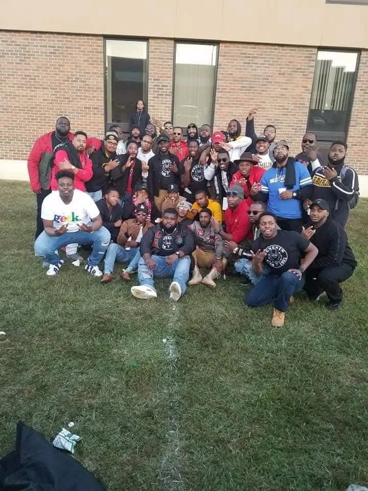 A large group of people posing outdoors on grass in front of a brick building with large windows. They are smiling, some making hand gestures, and appear to be at a social gathering or event.
