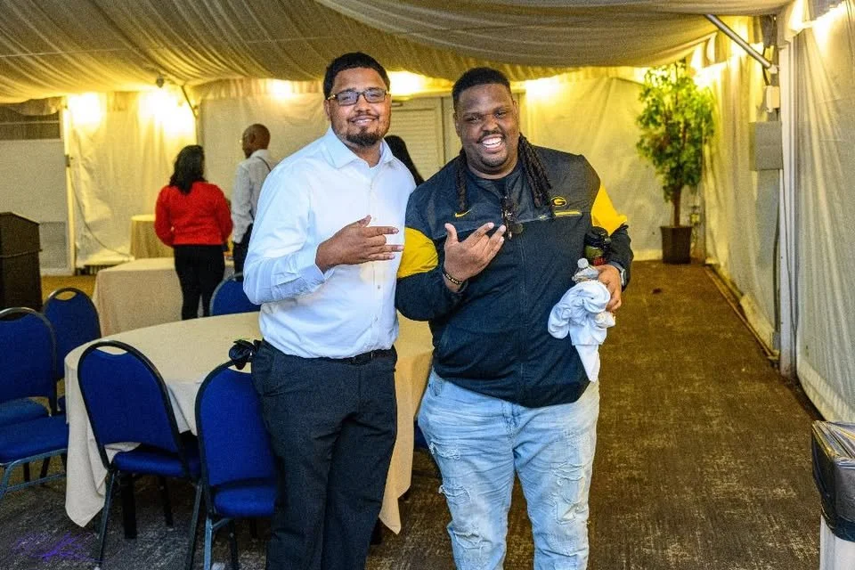 Two men posing and smiling inside a decorated event tent. One man is wearing glasses, a white shirt, and black pants; the other is dressed in a jacket and light-colored pants. There are tables and chairs in the background, with some other people pres