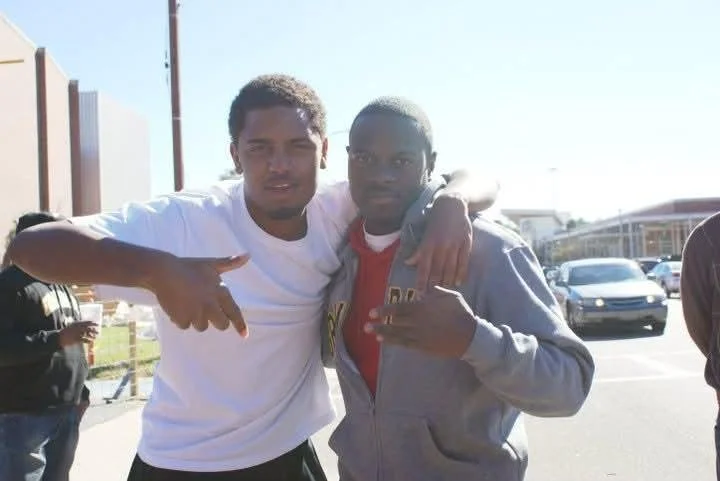 Two young men standing outdoors in a parking lot, with other people and cars in the background. The man on the left is wearing a white shirt and has his arm around the shoulder of the man on the right, who is wearing a gray hoodie and red shirt. Both