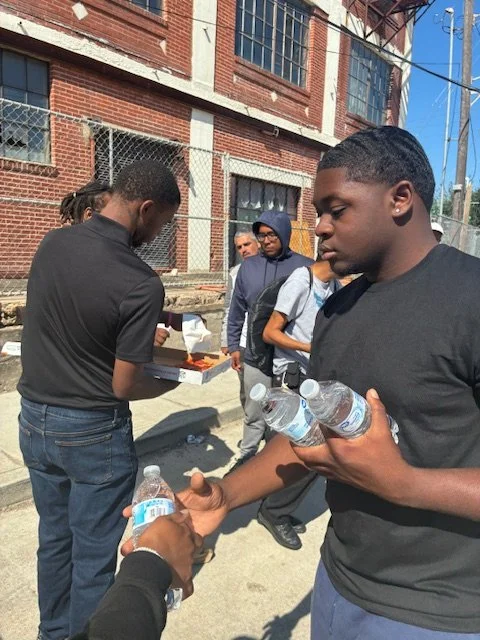 Group of people outdoors, with a city building in the background. One young man in the foreground holds several bottles of water, while others gather around.