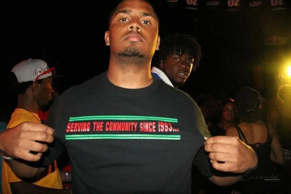 Young man wearing a black T-shirt with red and green text, showing his muscles at a social event or party with other people in the background.