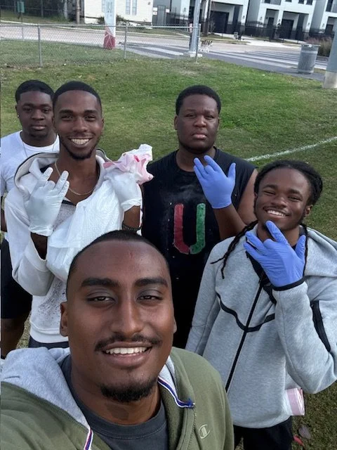 A group of five young men of African descent taking a selfie outdoors in a park, with some of them wearing gloves and smiling at the camera.
