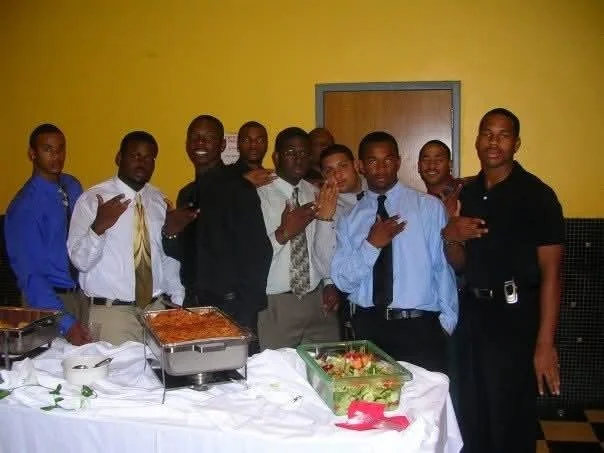 Group of nine men at a celebration with food, wearing formal and semi-formal clothing, standing in front of a yellow wall with a closed door.