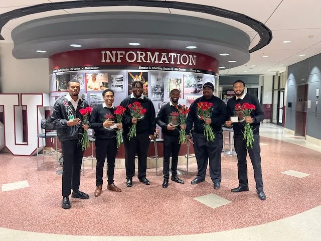 Six men standing in front of an information desk, holding bouquets of red roses, inside a building lobby.
