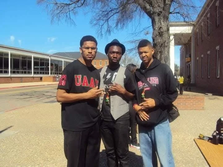 Three young men standing outside near a tree and brick building, posing for a photo.
