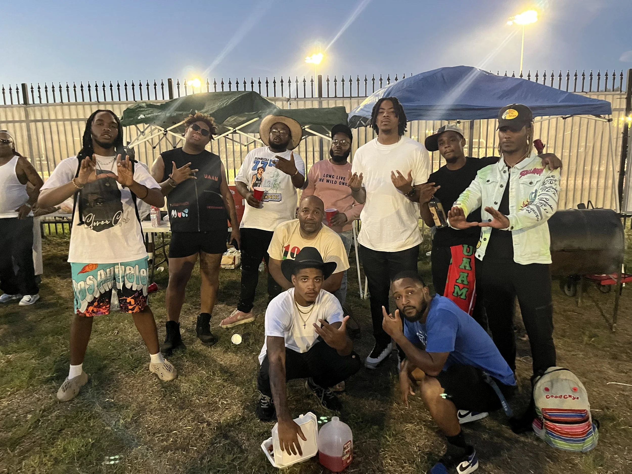 A group of eleven men gathered outdoors at night, some standing and some kneeling, in front of tables and tents, with a metal fence and bright streetlights in the background, enjoying a social event or party.