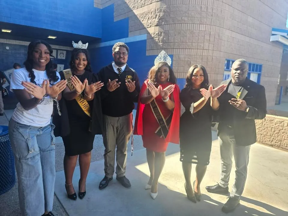 Group of seven people standing outside, some wearing crowns and sashes, celebrating at a school or event.