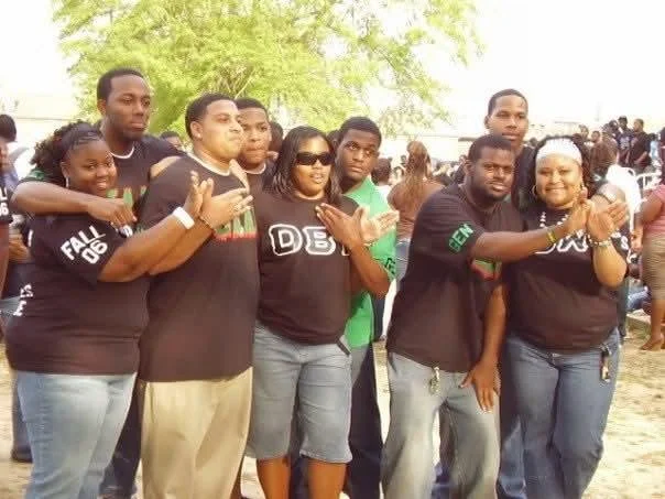 Group of eight people standing outdoors, some making hand gestures, with trees and other people in the background.