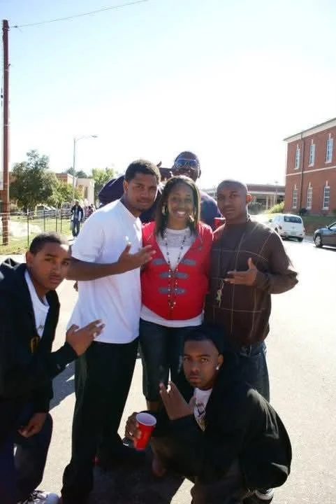A group of six young people posing outdoors on a street in a small town, with some vehicles and brick buildings in the background. They are making gestures with their hands and smiling.