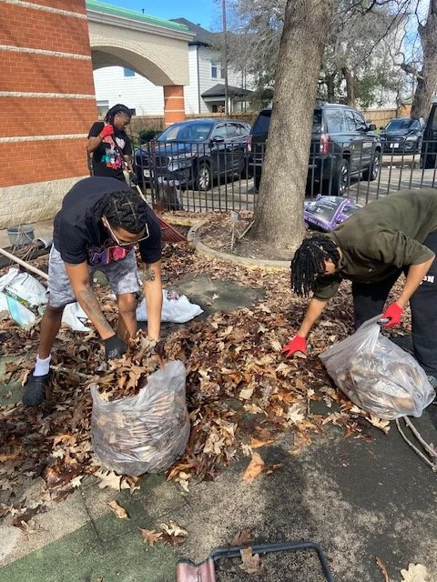 Two people raking and collecting fallen leaves into large trash bags in a parking lot, with a tree, a metal fence, and parked cars in the background.