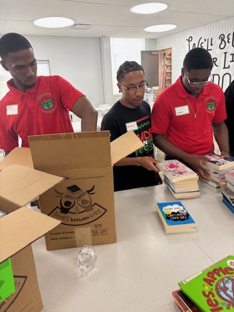 Three young men, two in red polo shirts and one in a black shirt, standing at a table organizing stacks of books in a library or book event space.
