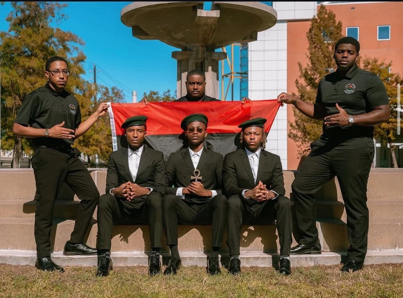 A group of five young Black men in black suits and berets sit and stand on steps outdoors, holding a red, black, and silver flag behind them during daytime. Two of the men stand on either side, holding the flag, while three sit in front, with the cen