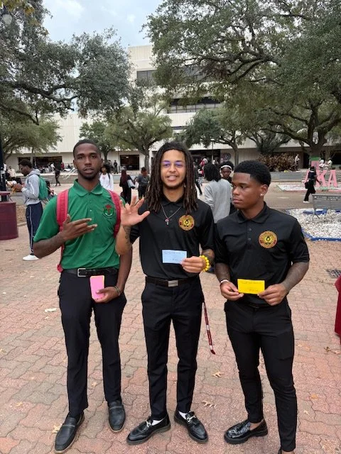 Three young men standing outdoors, two holding colorful cards, one making a hand gesture, with trees and a building in the background.