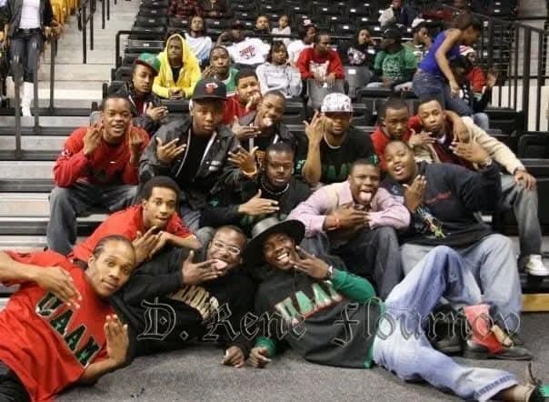Group of young African American men and boys sitting and lying in bleachers at an indoor sports arena, some making hand gestures, smiling, and posing for the camera.