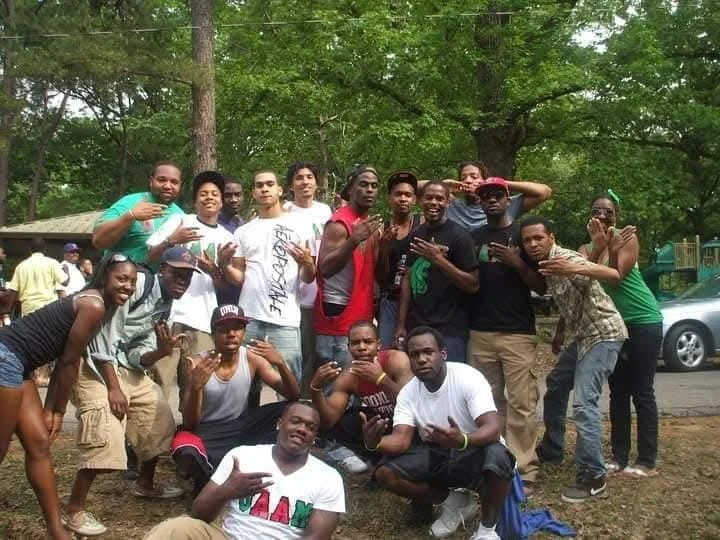 Group of young people outdoors in a park with trees, posing for a photo. Some making hand gestures, others smiling or striking poses.
