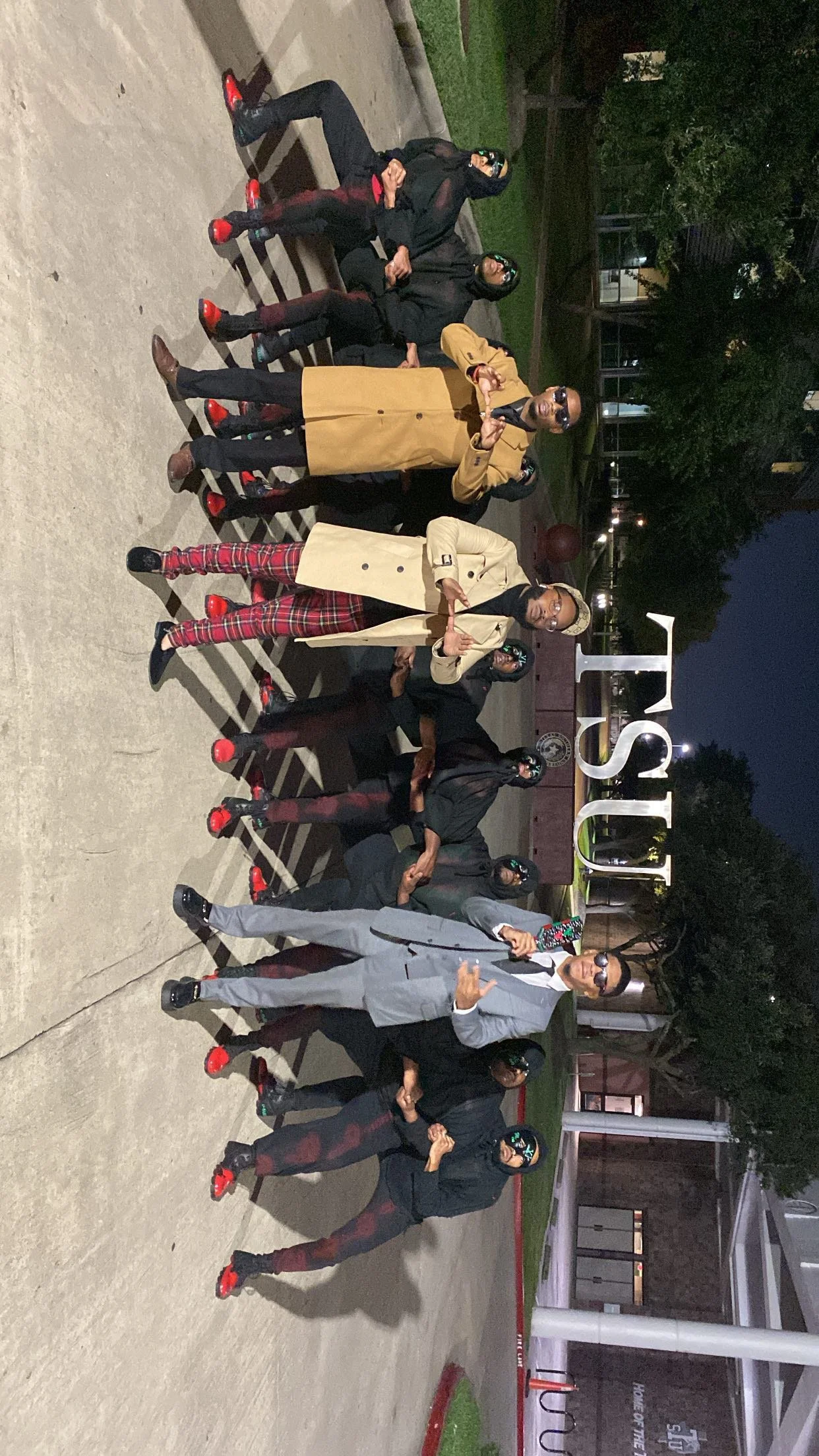 A group of people, most wearing black jackets and sunglasses, posing for a photo at night outside a building with a sign that reads 'TSU'.