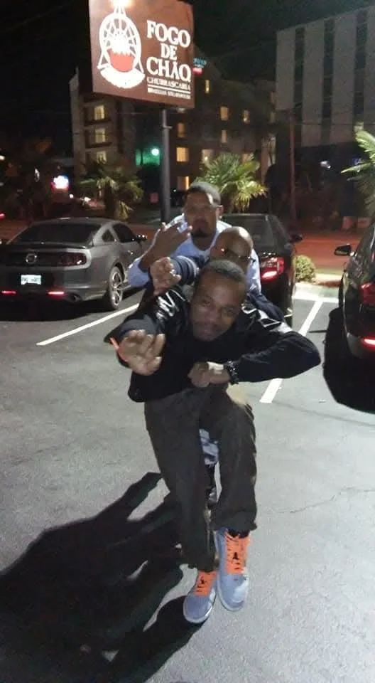 Two men at night in a parking lot with cars and palm trees, one carrying the other on his back. In the background, a sign reads 'Foco de Chao'.
