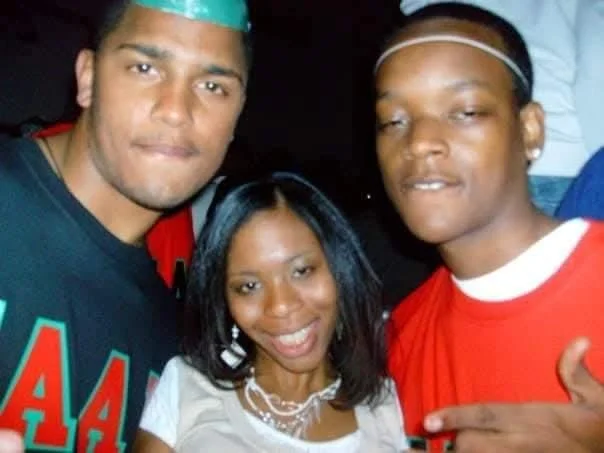 Three friends smiling and posing together at a party or gathering, two men and one woman, with casual clothing and accessories.