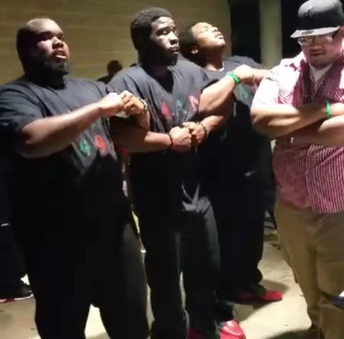 Four young men standing in a line with arms folded or clenched, wearing black and red shirts, participating in a group activity or demonstration.