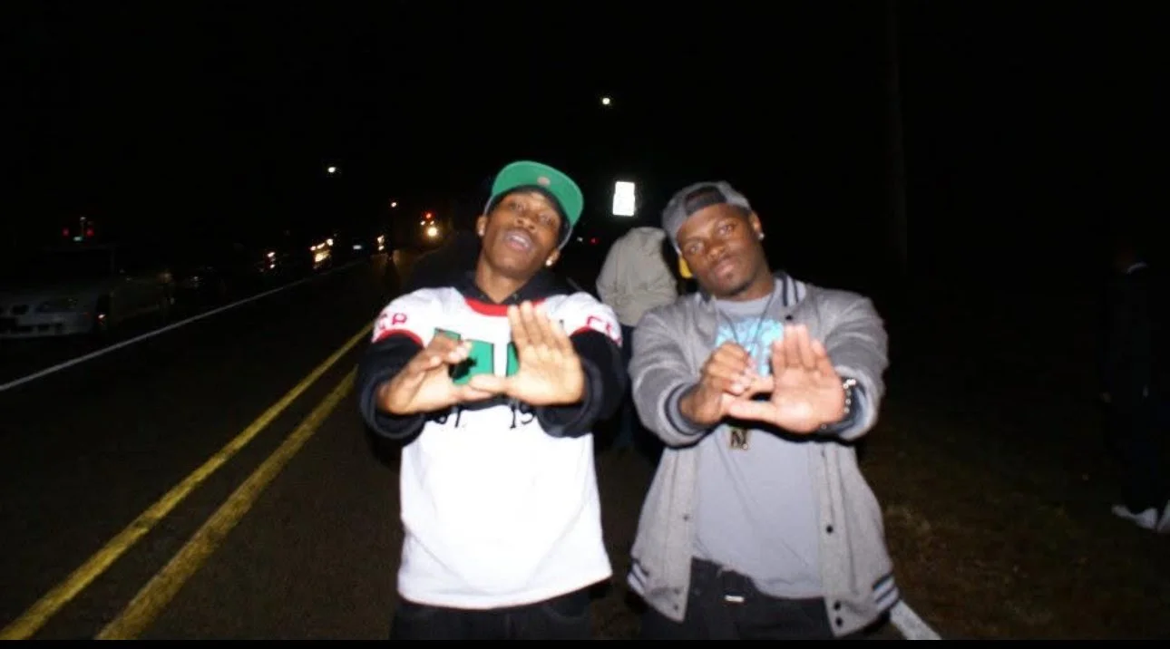 Two young men standing on a dark street at night, making hand gestures toward the camera.
