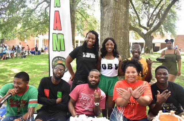 Group of people outdoors at a celebration or event, with some seated at a table and others standing behind, in front of a banner that partially reads 'AFR' and 'M.'