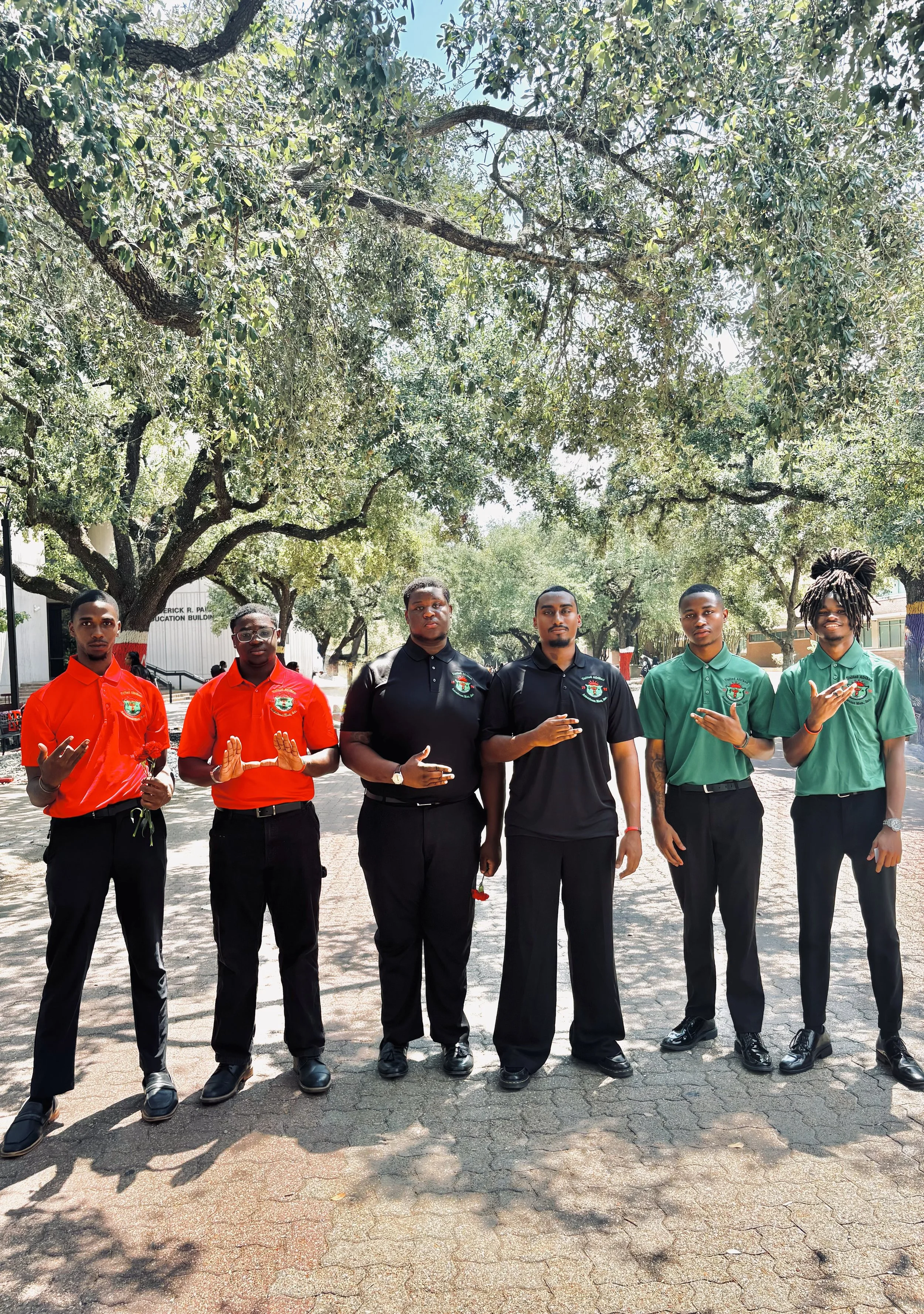 Group of six young people standing outdoors under trees, dressed in matching uniforms of red, black, and green shirts, and making hand gestures for a photo.