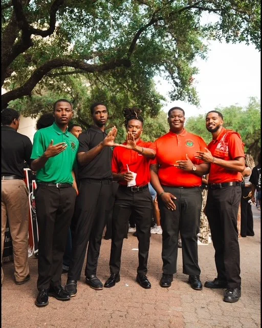 Group of five young adults standing outdoors under trees, wearing casual shirts in green, black, and red, posing together for a photo.