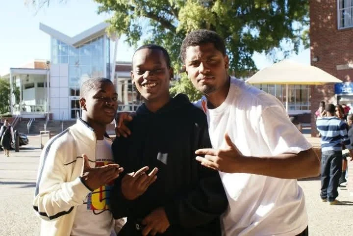 Three young men standing outdoors, smiling, with trees and a building in the background, some other people and an umbrella visible.